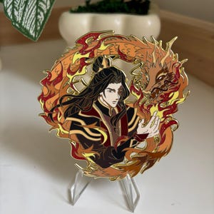 May include: A circular gold-toned enamel pin featuring a character with long dark hair and a scar over one eye, dressed in dark red and black robes. The character is surrounded by a swirling orange and red dragon made of glittery flames.