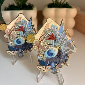 May include: Two gold-toned metal enamel pins featuring a circular design with a central globe, city buildings, a red cape, and the text Daily Planet and Hall of Justice. The pins are displayed on clear plastic stands, showcasing intricate details.