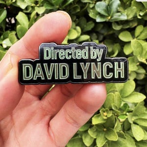 May include: A black enamel pin featuring the text "Directed by DAVID LYNCH" in green. The rectangular pin has a black border. It is held in a hand, set against a backdrop of green foliage.
