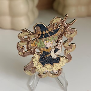 以下が含まれることがあります： 淡い緑色の髪をしたキャラクターが、模様入りの紺色の魔女の帽子とドレス、黄色いフリルのスカートを身につけた金色のエナメルピン。キャラクターは小さな白い生き物を抱えており、周囲は茶色のリボンで囲まれている。