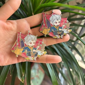 May include: Two enamel pins depicting a cartoon character with white hair, red eyes, and a blue and gold outfit. The pins are set against a red and gold background with a tent-like structure. The character is seated on a cloud with a small animal.