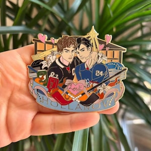 May include: A gold-colored enamel pin featuring two male figures embracing, set against a hockey rink backdrop with pink hearts. The pin includes hockey sticks, a ribbon with the word "HOLLANOV," and a heart with "Love."