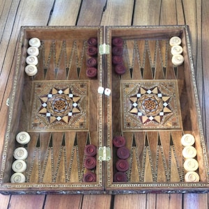 Puede incluir: Un tablero de backgammon de madera abierto, listo para jugar. El tablero presenta un diseño de estrella con incrustaciones decorativas en el centro, con triángulos claros y oscuros alternados. Incluye fichas blancas y granates y dados.