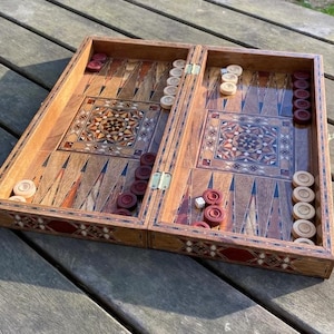 Puede incluir: Un tablero de backgammon de madera abierto con patrones decorativos incrustados. La superficie de juego presenta triángulos claros y oscuros alternados. Las fichas rojas y blancas están dispuestas en el tablero, junto con dados. El tablero está sobre una mesa de madera.