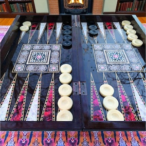 Pode incluir: Um tabuleiro de gamão de madeira escura com peças de jogo em marfim e preto. O tabuleiro apresenta um design decorativo embutido com detalhes em vermelho, rosa e branco. O tabuleiro está aberto e pronto para jogar.