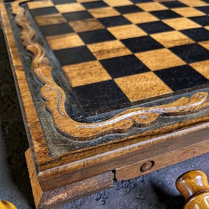 Peut inclure: Gros plan sur un échiquier en bois avec une bordure sculptée. Le plateau présente des cases noires et marron clair alternées. La bordure est ornée d'un motif détaillé. Le bois a une finition polie, pour un aspect classique et élégant.