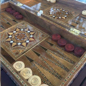 以下が含まれることがあります： 星模様と幾何学模様が象嵌された木製のバックギャモンボード。ボードには赤と白のゲームピース、サイコロ、装飾的なボーダーが特徴です。ゲームボードは木製で、研磨された仕上げが施されています。