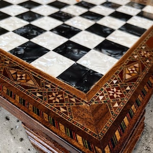 May include: Close-up of a handcrafted chess board. The playing surface features alternating black and white squares. The wooden frame is intricately inlaid with geometric patterns in various colours, showcasing detailed craftsmanship.
