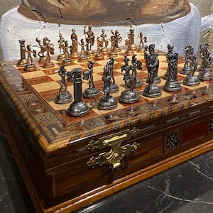 Puede incluir: Un juego de ajedrez de madera con piezas de ajedrez de metal talladas intrincadamente. El tablero de ajedrez está incrustado con un patrón de madera clara y oscura. Las piezas de ajedrez son en tonos plateados y bronce, que representan figuras en varias poses. El juego se exhibe sobre una superficie oscura.