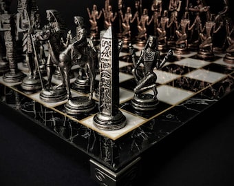 May include: An Egyptian-themed chess set with silver and bronze pieces on a black and white checkered board. The chess pieces include pharaohs, obelisks, and figures riding camels. The board has a black marble-like border.
