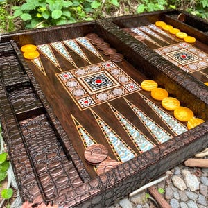 Puede incluir: Un detallado tablero de backgammon con una superficie de juego de madera marrón oscuro. El tablero presenta patrones incrustados con diseños geométricos y acentos coloridos. Las fichas marrones y amarillas están dispuestas en el tablero, listas para jugar. El marco exterior tiene un patrón texturizado similar al de un cocodrilo.