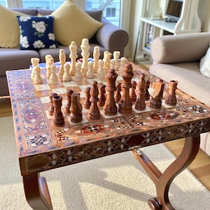 Puede incluir: Un juego de ajedrez de madera con un tablero decorativo con incrustaciones. Las piezas del ajedrez son de color marrón claro y oscuro. El tablero está sobre una mesa de madera con patrones incrustados. La mesa mide unos 76 cm x 76 cm.