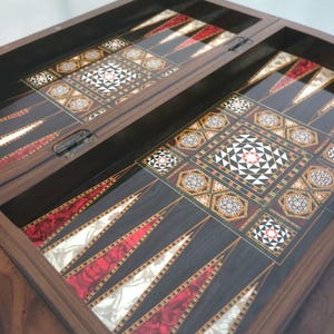 May include: Close-up of a backgammon board. The dark wood board features inlaid geometric patterns in red, white, and gold. The playing surface has alternating red and white triangles and decorative central squares.