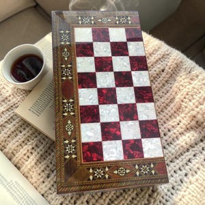 Puede incluir: Un tablero de ajedrez decorativo de madera con una superficie de juego a cuadros rojos y blancos. El tablero está enmarcado con patrones incrustados ornamentados y descansa sobre una superficie suave y texturizada. Una taza de té y un libro están en el fondo.