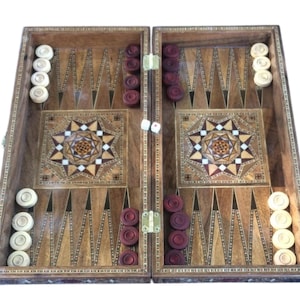 Puede incluir: Un juego de backgammon de madera con diseños de estrella incrustados. El tablero está abierto, mostrando fichas blancas y granates, y dados. El tablero es de madera con un borde decorativo.