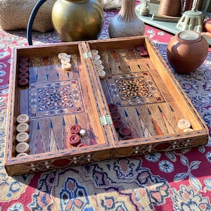 Puede incluir: Un juego de backgammon de madera con patrones decorativos incrustados. El tablero de juego está abierto, revelando fichas rojas y blancas. El juego está sobre una alfombra estampada, con otros objetos decorativos en el fondo.