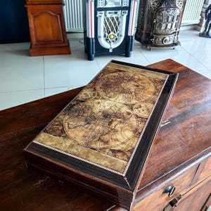 Könnte beinhalten: Eine rechteckige Holzkiste mit einem Weltkarten-Design auf dem Deckel. Die Karte ist in Braun- und Goldtönen gehalten und weist detaillierte Elemente auf. Die Kiste hat einen dunkelbraunen Rahmen und steht auf einer Holzoberfläche.