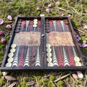 Könnte beinhalten: Ein offenes Backgammon-Spielbrett aus Holz mit dunkelbraunen und roten dreieckigen Punkten. Das Spielbrett hat helle und dunkle braune Spielsteine und Würfel. Das Spiel liegt auf grünem Moos und verstreuten rosa Blütenblättern. Das Brett misst ca. 40 cm.