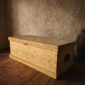 May include: A rectangular wooden chest in a light brown hue. It features a hinged lid, a small keyhole, and a metal handle. The chest is positioned on a wooden floor, showcasing its aged appearance.