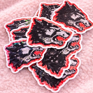 Op de afbeelding: Een verzameling stickers met een grommend wolfskopontwerp. De wolf is zwart en wit met een rode omtrek, en de stickers hebben een witte rand. De stickers liggen verspreid op een roze, getextureerd oppervlak.