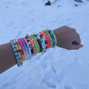 May include: A selection of colourful, beaded bracelets stacked on a wrist. The bracelets feature a variety of colours including pink, blue, orange, and yellow. Some bracelets have gold accents and a star charm. The background is a snowy landscape.