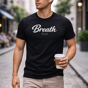 May include: Black t-shirt with the word "Breath" in white script, and Japanese characters below. The person is wearing khaki pants and holding a coffee cup. The background is a city street.