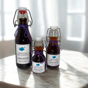 Könnte beinhalten: Drei Glasflaschen Madagaskar-Vanilleextrakt mit Bügelverschlüssen und lila Bindfäden. Die Flaschen sind unterschiedlich groß und tragen jeweils ein Etikett mit der Aufschrift "Madagascar Vanilla Extract". Der Extrakt hat eine dunkelbernsteinfarbene Farbe.