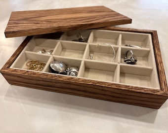 May include: A wooden jewelry box with a hinged lid, featuring a light-colored interior divided into compartments. The box is filled with various pieces of jewelry, including necklaces, rings, and earrings. The wood has a natural grain pattern.