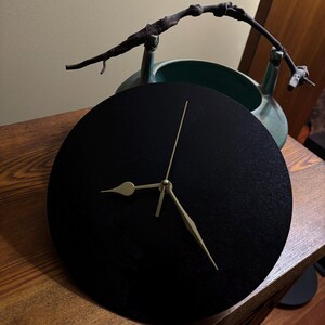 May include: A circular black wall clock with a textured surface and thin gold-coloured hands. The clock face lacks numbers or markers. Behind the clock sits a teal ceramic vessel with a dark brown, gnarled wooden branch handle attached.