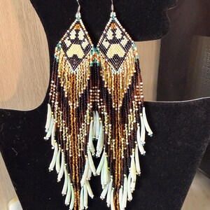 Peut inclure: Une paire de longues boucles d'oreilles perlées. Chaque boucle d'oreille présente un motif en forme de losange avec un motif de pattes de couleur crème et noir. Les boucles d'oreilles ont de longues franges perlées dans les tons de marron, d'or et de blanc, avec de petits accents turquoise.