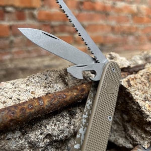 Puede incluir: Una navaja multiusos con mango color beige y múltiples hojas, incluyendo una sierra y un abrebotellas. Las hojas son plateadas y el mango presenta un agarre texturizado y un emblema de escudo. La navaja está parcialmente abierta.