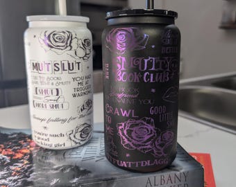 May include: Two insulated tumblers with straws, one white and one black, each adorned with floral designs and text. The white tumbler reads "MUT SLUT" and the black one says "SMUTTY BOOK CLUB". The tumblers rest on a stack of books.