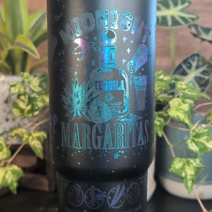Op de afbeelding: Zwarte geïsoleerde beker met een teal en iriserend ontwerp. De beker heeft de woorden "Midnight Margaritas" met illustraties van een tequila fles, cocktail en botanische elementen. De onderkant van de beker heeft extra tekst.