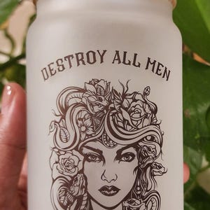 Peut inclure: Gobelet en verre givré en forme de canette avec un couvercle en bambou et une paille en verre transparent. Le gobelet présente une illustration de Méduse avec le texte "DESTROY ALL MEN WHO ABUSE THEIR POWER" en noir.