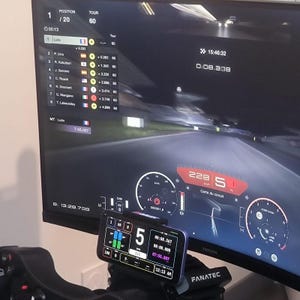 Peut inclure: Configuration de simulateur de course avec un grand écran affichant un circuit et des données de voiture. Un smartphone affiche des informations supplémentaires. L'écran affiche les temps au tour, les positions et la vitesse. Le setup comprend un volant et le logo "FANATEC".