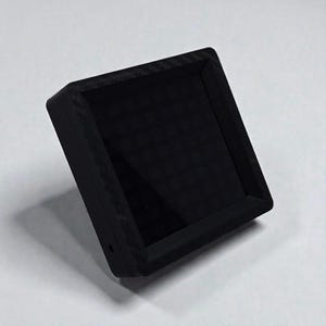 Peut inclure: Petit appareil électronique noir de forme carrée avec un écran. L'appareil présente un motif texturé semblable à de la fibre de carbone sur les bords. L'écran est sombre et l'appareil est légèrement incliné.