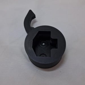 Puede incluir: Una herramienta negra, impresa en 3D, con una base circular y un asa. La base tiene un interior hexagonal con un agujero central y un recorte en forma de cruz. El asa se curva hacia arriba.
