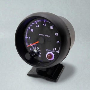 Może przedstawiać: Czarny obrotomierz samochodowy z okrągłą tarczą. Wskaźnik pokazuje RPM x1000, ze wskazówką wskazującą aktualne obroty. Posiada lampkę zmiany biegów i pokrętło ustawień. Cyfry są białe, a wskazówka pomarańczowa.
