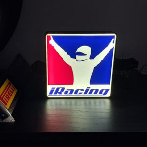 May include: Illuminated square sign with the iRacing logo. The sign features a white silhouette of a race car driver with arms raised against a red and blue background. The iRacing text is in white.