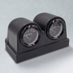 May include: Black dual gauge instrument with two circular dials. One dial reads TEMP/°C, the other reads OIL/°C. The temperature gauge shows 120 degrees, and the oil gauge shows 150 degrees. The gauges are housed in a black plastic casing.