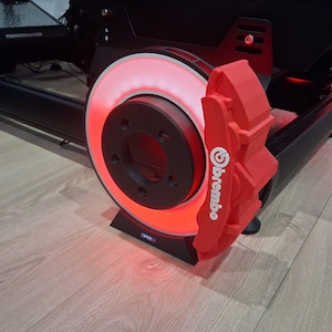May include: A red Brembo brake caliper and rotor assembly with a glowing red light. The assembly is part of a racing simulator setup. The Brembo logo is visible on the caliper.