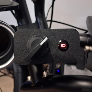 May include: A black control panel featuring a rotary dial and a red illuminated button. The dial has a white indicator line. The button has a battery symbol. The panel is mounted on a black frame.