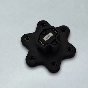 May include: A black, star-shaped component with a central, rectangular module. The module has a barcode sticker. The star has six circular holes and is on a white surface. The component appears to be made of plastic.