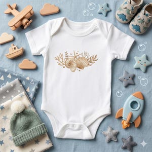 Peut inclure: Body de bébé blanc avec un motif de coquillages brodés en or. Le body est entouré de jouets pour bébés, dont un avion, un bateau et des nuages en bois, ainsi que des jouets en forme d'étoiles et des chaussures de bébé. Thème nautique.