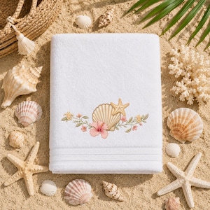 Könnte beinhalten: Ein weißes, rechteckiges Badetuch mit einer Stickerei in der Mitte, die eine beige Jakobsmuschel, einen goldenen Seestern, eine rosa Hibiskusblüte und grüne Ranken mit kleinen rosa Blüten zeigt, die horizontal angeordnet sind.
