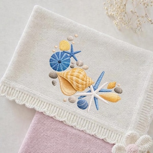 May include: Cream knitted blanket featuring an embroidered seashell design. The design includes a large tan seashell, blue starfish, and small yellow shells. The blanket has a scalloped edge and is folded over a pink blanket.