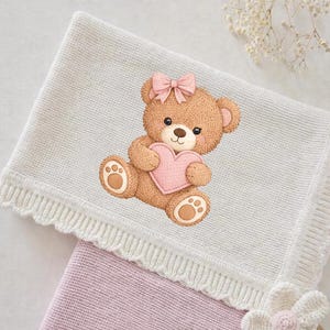 Può includere: Una coperta per neonati in maglia grigio chiaro con bordo smerlato e un orsacchiotto marrone che tiene un cuore rosa. L'orsacchiotto ha un fiocco rosa. Sotto è visibile una seconda coperta rosa.