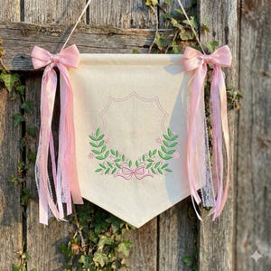 May include: A cream-colored fabric wall banner featuring an embroidered green leafy wreath with small pink flowers and a central pink bow. The banner is suspended by a string and decorated with long, flowing light pink satin and lace ribbons.