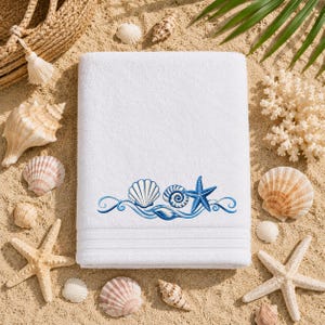 May include: A white rectangular bath towel featuring an embroidered design of a blue scallop shell, a spiral shell, and a starfish connected by swirling blue lines. The towel has a textured fabric surface and horizontal decorative bands.