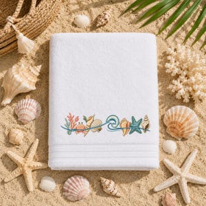 May include: A white rectangular bath towel featuring an embroidered horizontal band of colorful marine life, including a small orange crab, various tan seashells, a blue starfish, and a stylized blue wave pattern across the center.